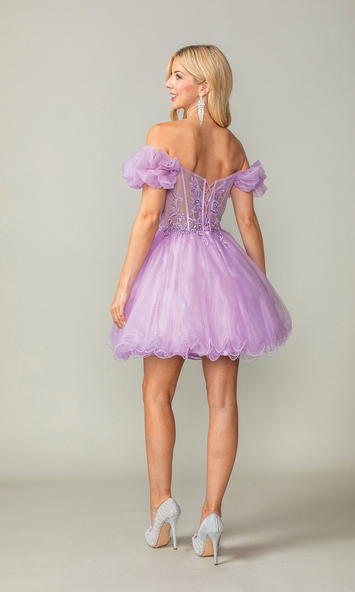 A woman faces away in the Ruffle-Sleeve Short Corset Homecoming Dress 3379, an off-the-shoulder lavender tulle design with puff sleeves and a corset bodice, paired with silver heels, set against a plain background.