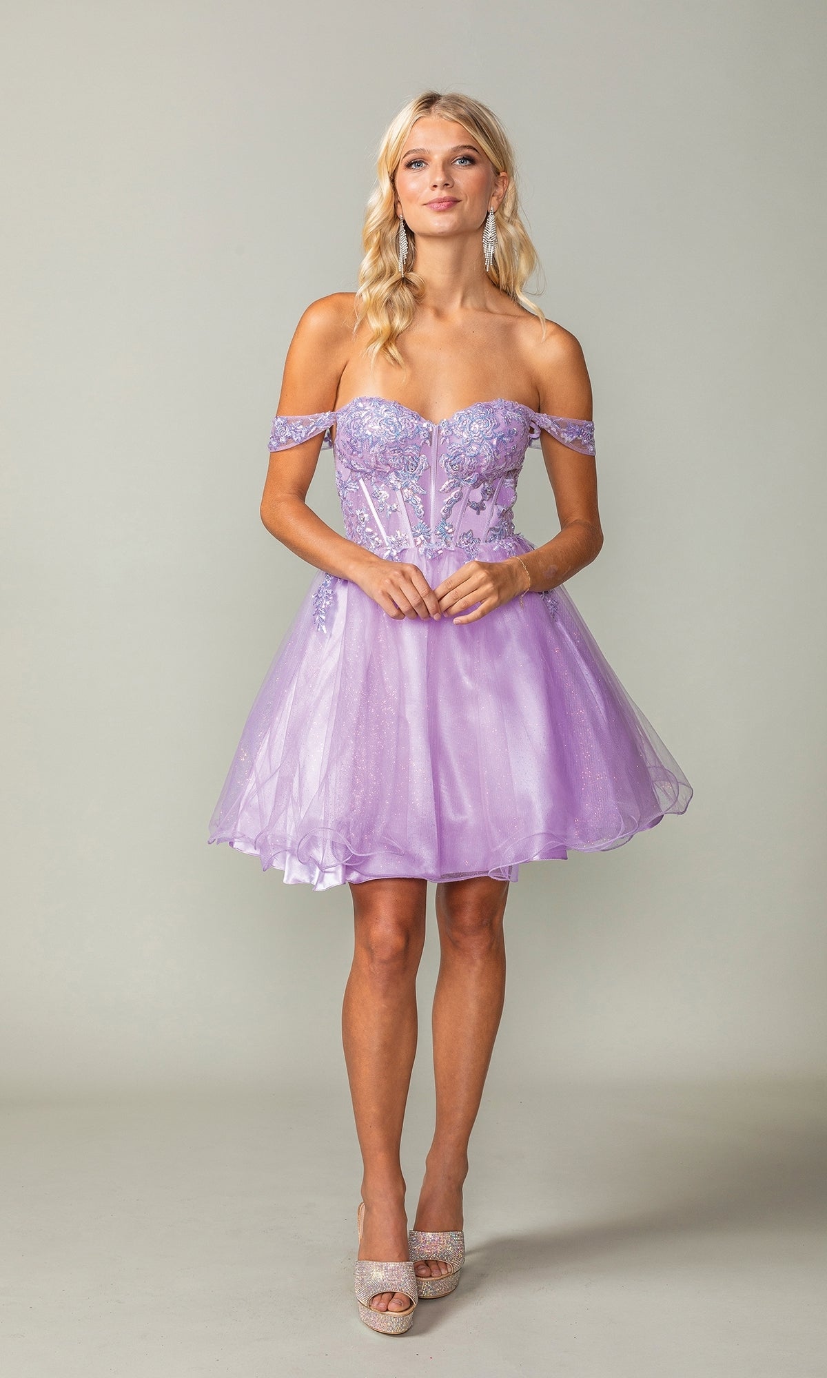 A woman poses in the Dancing Queen 3383 short corset homecoming dress, featuring floral embellishments and a sparkly sheer bodice, styled with high heels against a plain background.