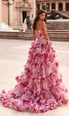 Wearing the La Femme 33842 long prom dress, a woman in a strapless sweetheart pink floral gown with a layered ruffle skirt stands outdoors by a fountain, glancing over her shoulder.