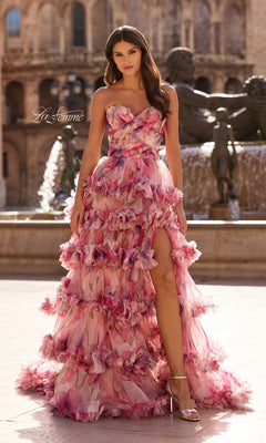 A woman in the La Femme 33842 long prom dress—a strapless pink floral gown with a sweetheart neckline, high slit, and layered ruffle skirt—stands outdoors by a stone fountain near historical buildings.