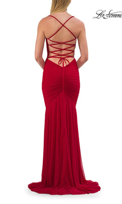 A woman is wearing the Strappy Long Net-Jersey Prom Dress: La Femme 33857 in bold red, which showcases a backless design with crisscross lacing and sophisticated ruched detailing, as she stands facing away from the camera.