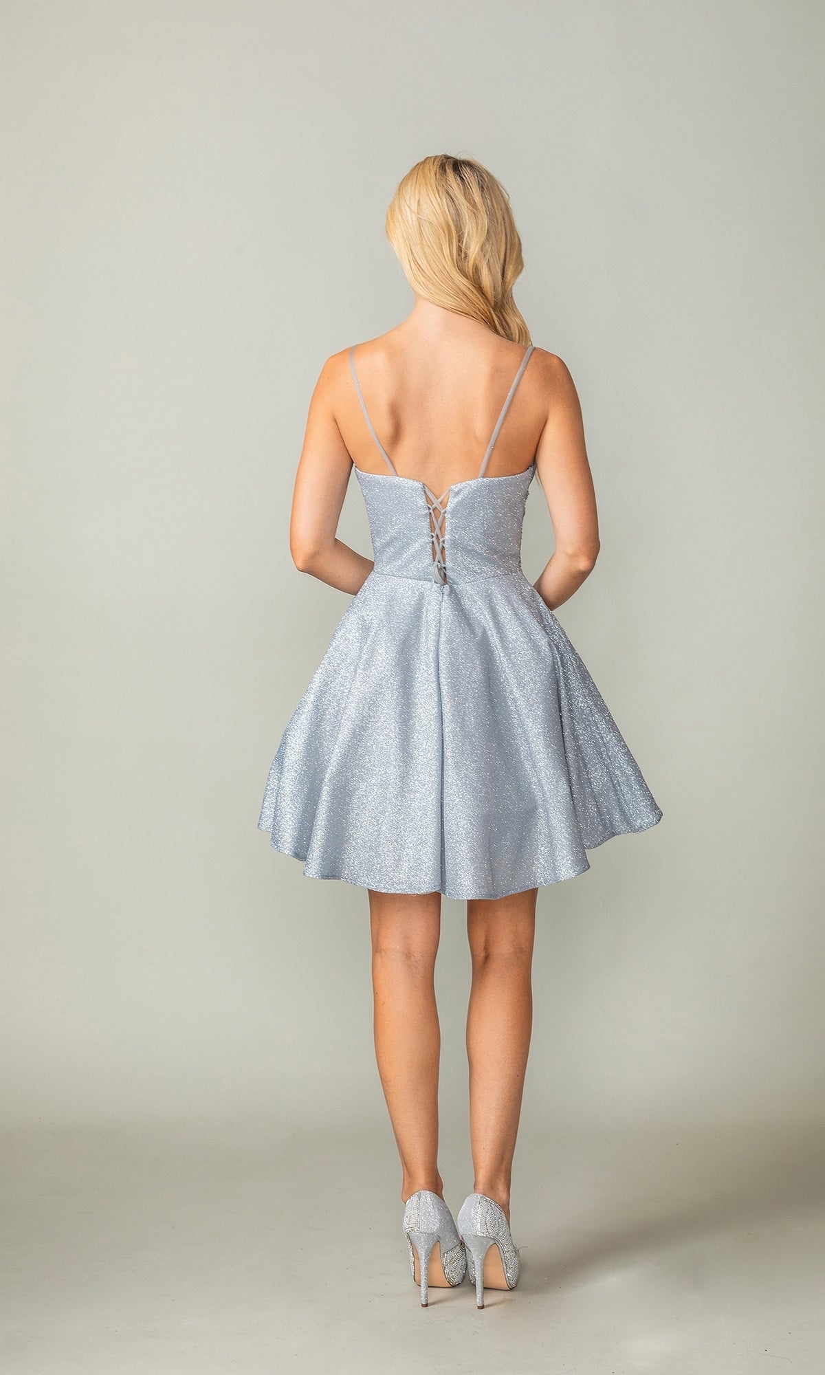 A woman faces away, wearing the Cowl-Neck Short Glitter Homecoming Dress 3386—a sparkly light blue, lace-up mini dress with thin straps—paired with matching high heels.