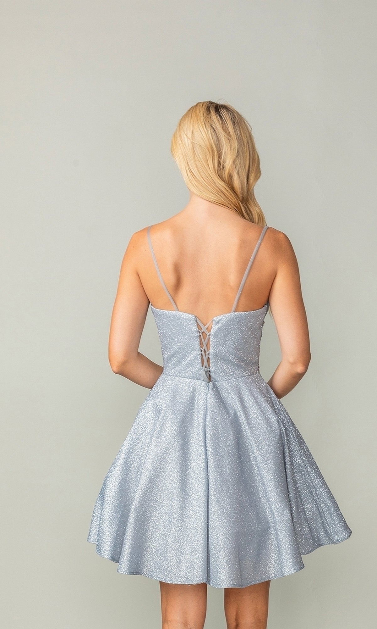 A woman with long blonde hair wears the Cowl-Neck Short Glitter Homecoming Dress 3386—a sparkly, lace-up cocktail dress with thin straps—standing against a plain light background.