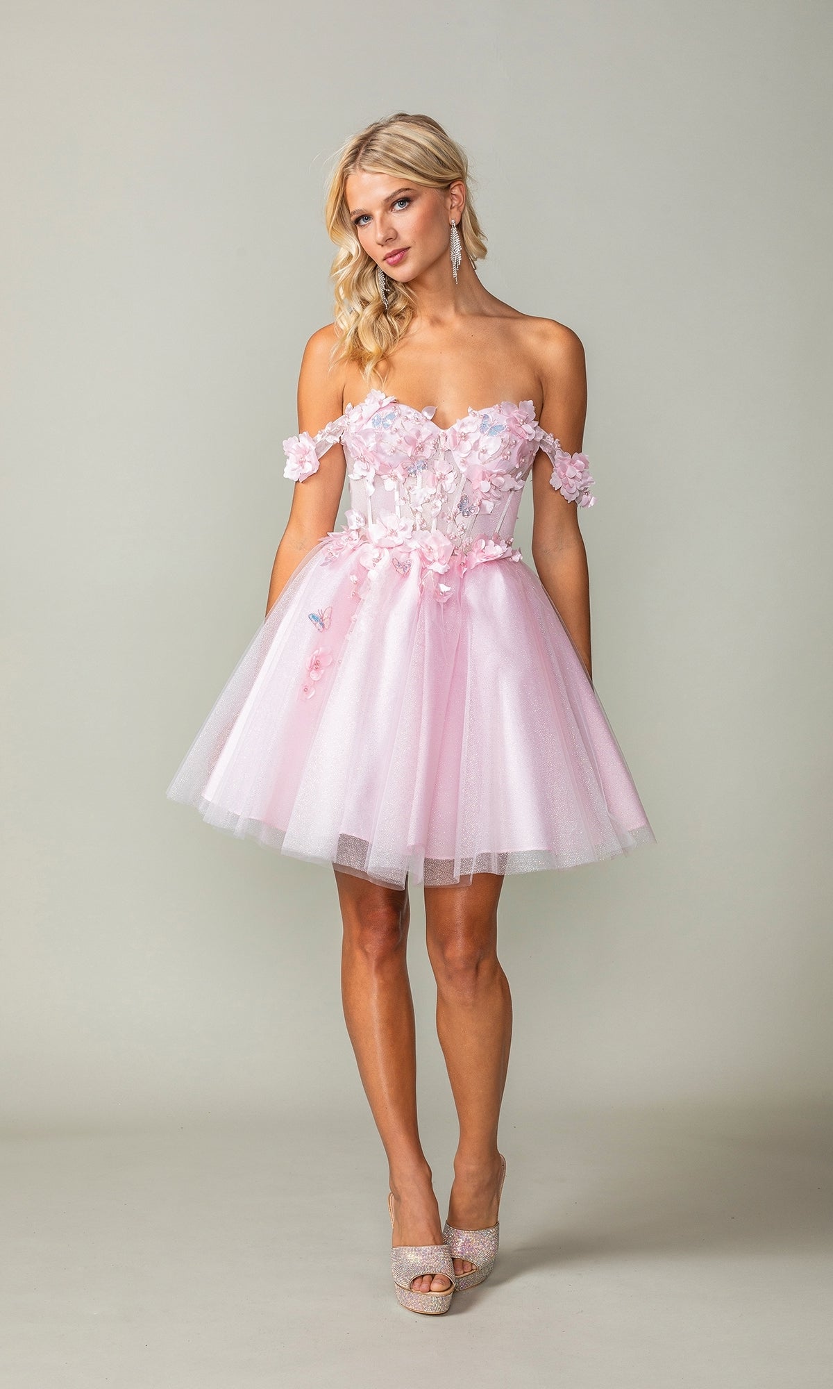 A woman models the Off-Shoulder Short Babydoll Homecoming Dress 3388, featuring floral details and a tulle skirt, paired with sparkly heels and posed against a plain background.