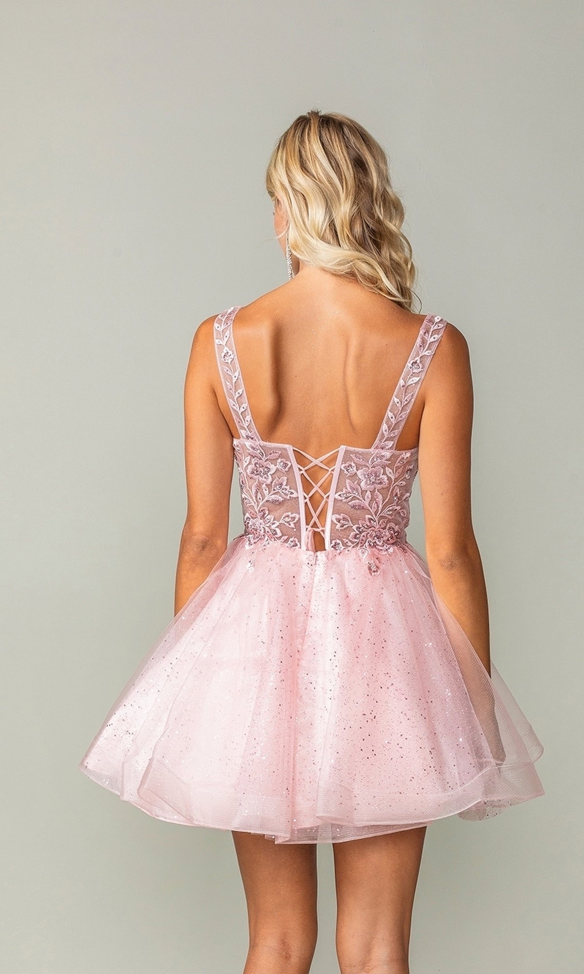 A woman with wavy blonde hair models the Embroidered-Bodice Short Lace-Up Hoco Dress 3390, featuring a flared glitter skirt and lace-up back, standing against a plain background.