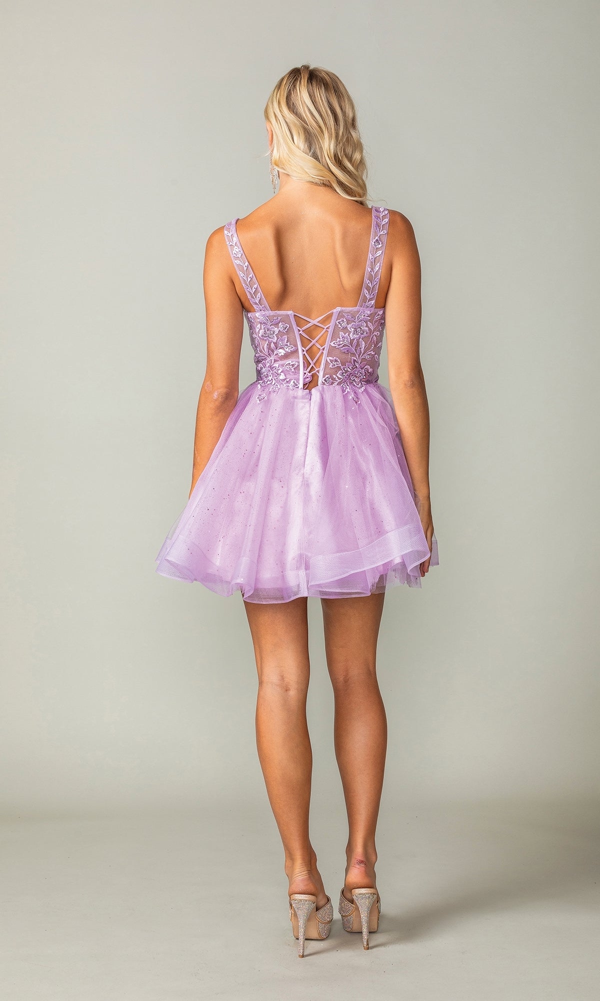 A woman faces away in the Embroidered-Bodice Short Lace-Up Hoco Dress 3390, a lavender babydoll dress with floral embroidery, lace-up back, and sheer layers. She pairs it with nude high heels.