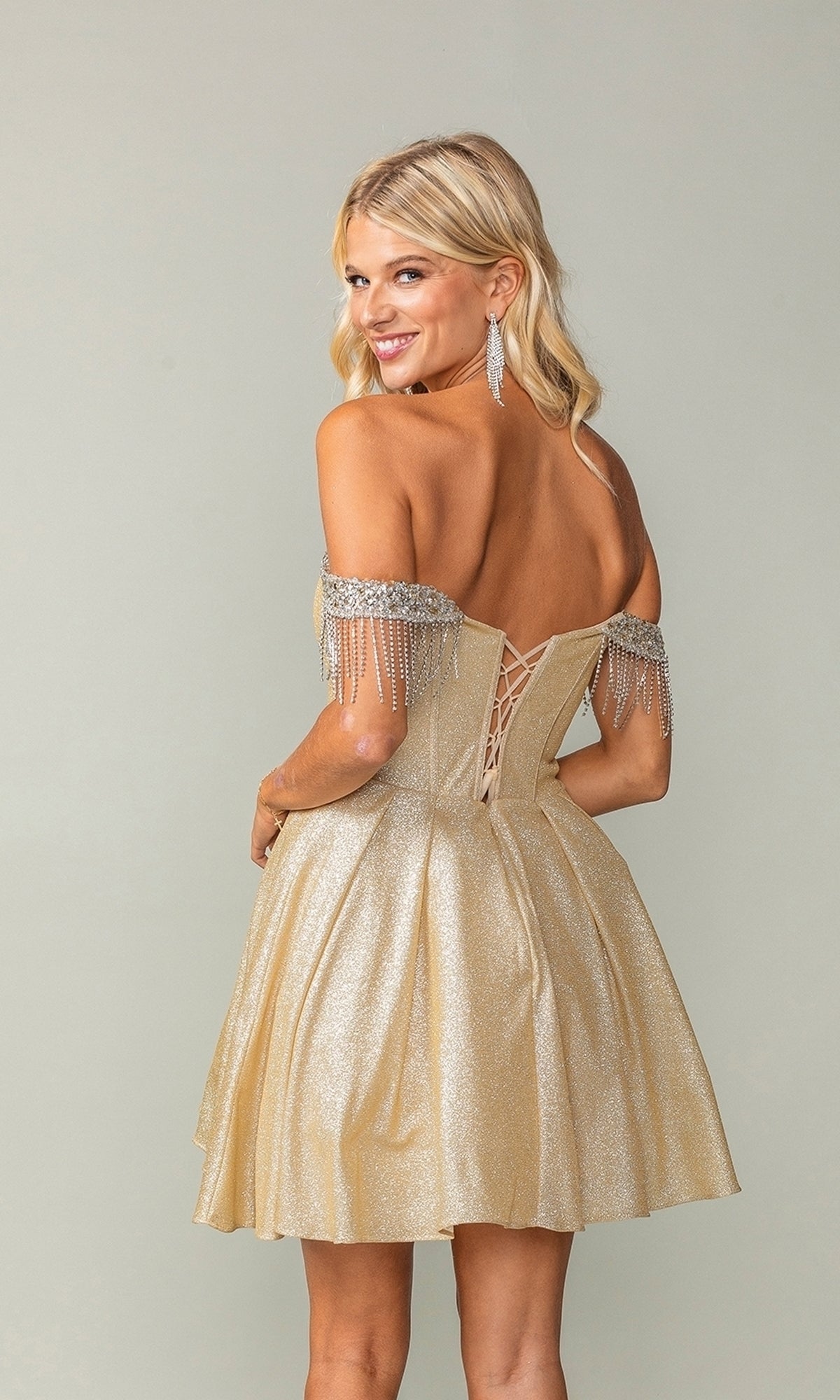 Smiling over her shoulder, a young woman with long blonde hair wears the Fringed Off-Shoulder Short Homecoming Dress 3392, featuring a sparkling corset bodice, beaded fringe accents, and lace-up back against a plain background.