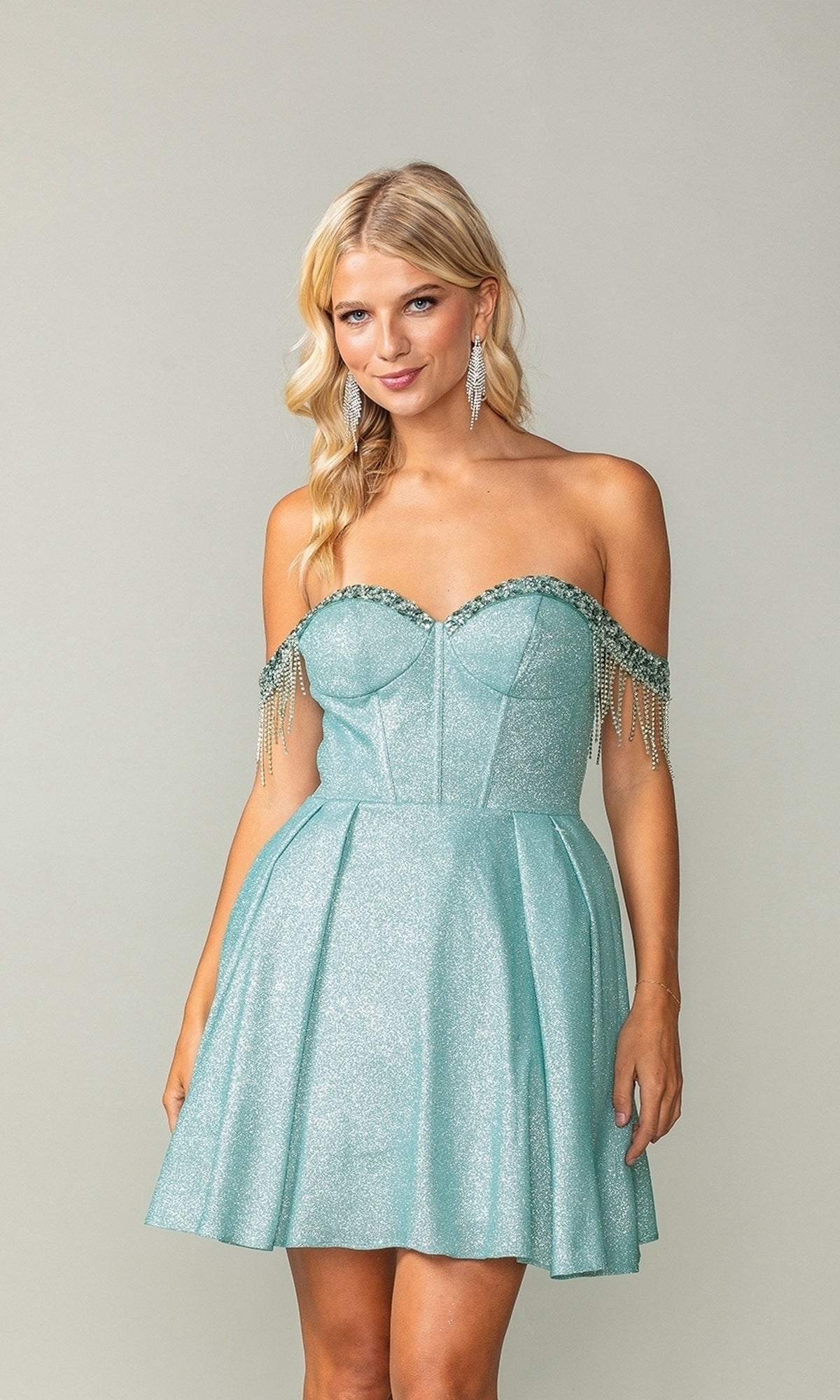 A woman in the Fringed Off-Shoulder Short Homecoming Dress 3392 with a corset bodice and beaded fringe stands against a plain background, her light blue glitter dress sparkling elegantly.