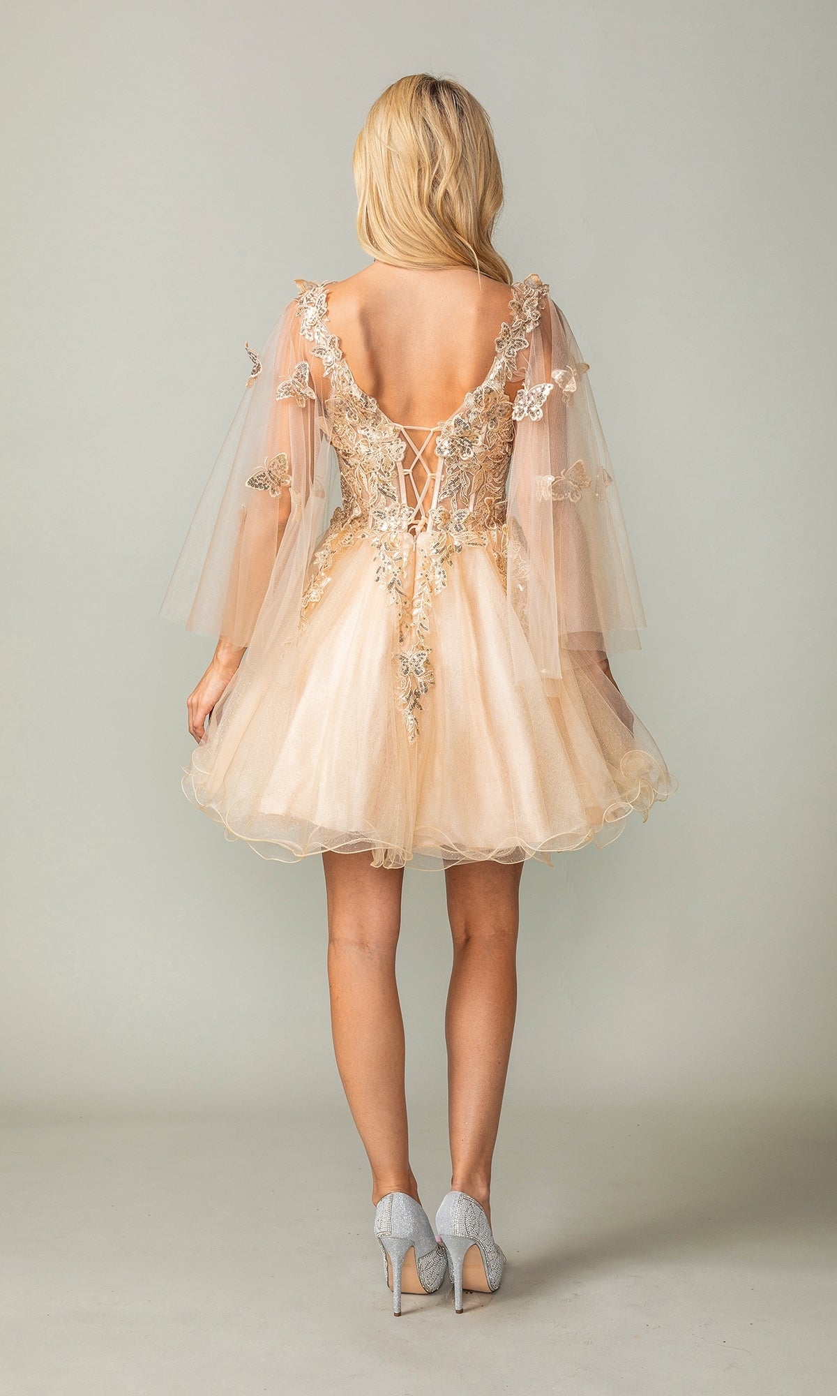A blonde woman models the Butterfly-Cape Short A-Line Homecoming Dress 3393 in light peach with sequin appliques, lace-up back, sheer sleeves, and grey heels, standing against a plain background.