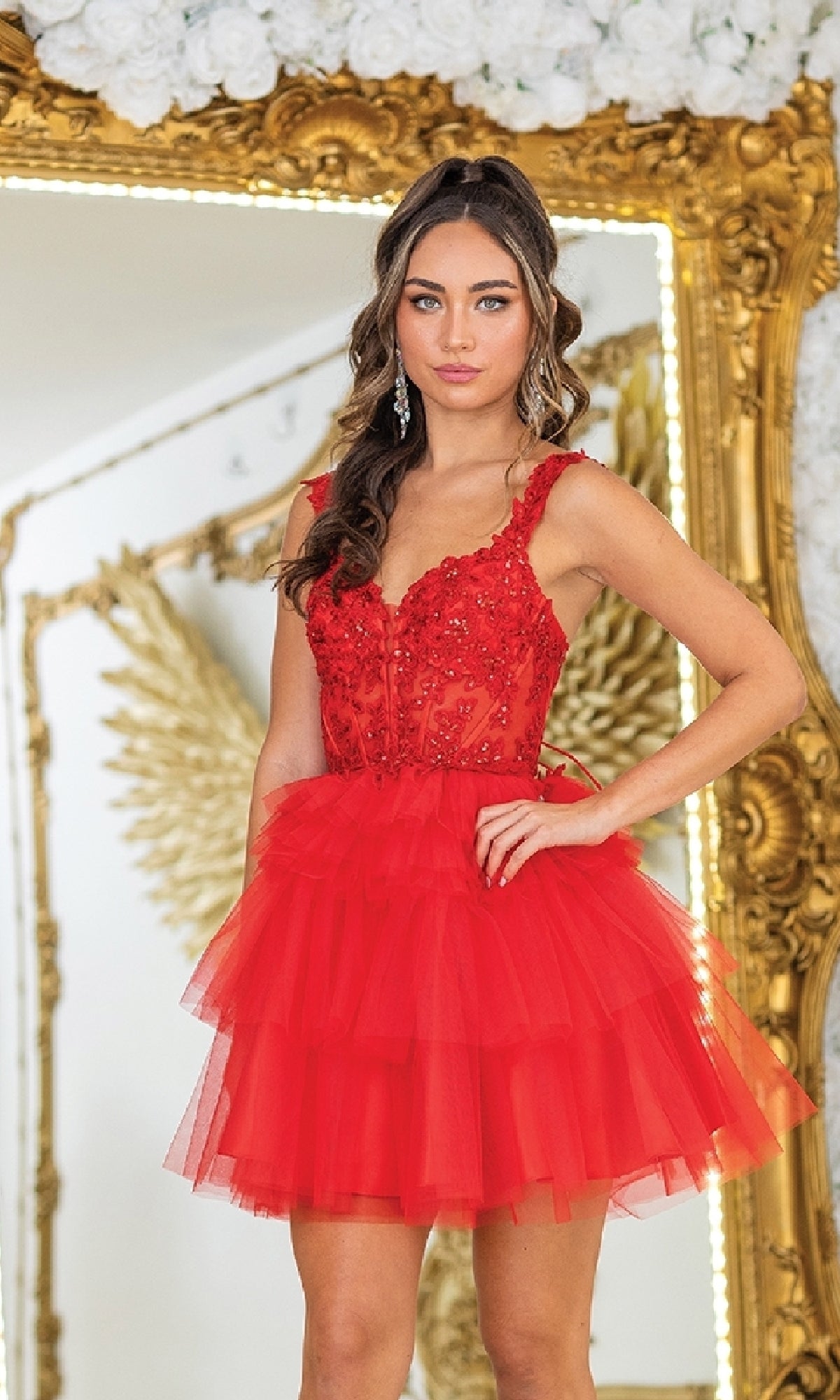 A woman stands before a gold-framed mirror wearing the Short Babydoll Prom Dress: Dancing Queen 3402, featuring floral embellishments and a sheer corset bodice. White flowers adorn the top of the mirror.