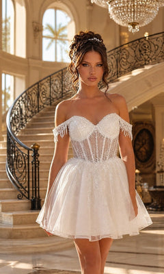 A woman wearing the Short Glitter Babydoll Prom Dress: Dancing Queen 3405 in Off White, featuring an off-the-shoulder sheer bodice, stands before a grand staircase in an elegant, well-lit interior.
