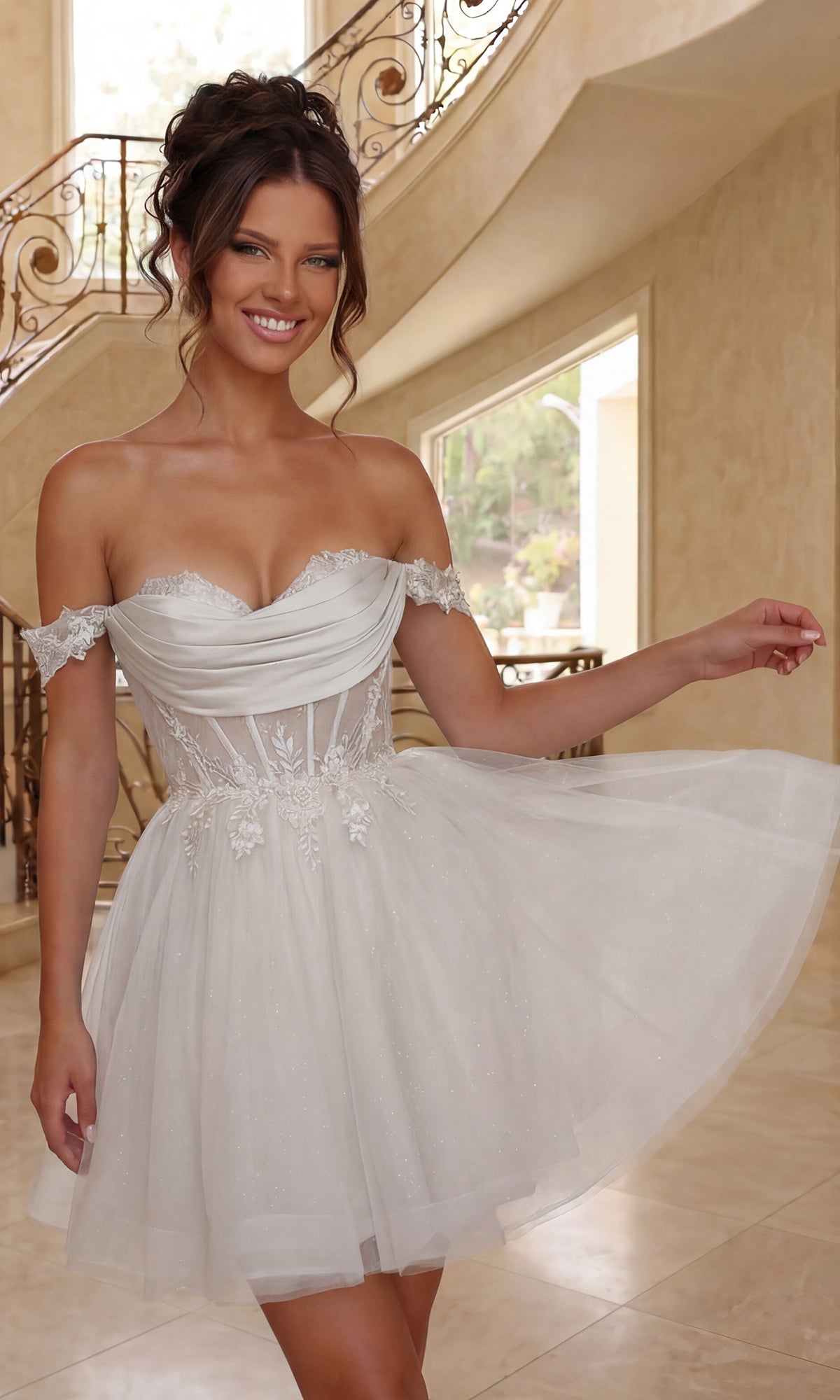 A woman in an off-shoulder, off white, short tulle Dancing Queen 3409 babydoll prom dress with floral details stands indoors, smiling and holding her skirt.