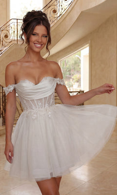 A woman in an off-shoulder, off white, short tulle Dancing Queen 3409 babydoll prom dress with floral details stands indoors, smiling and holding her skirt.