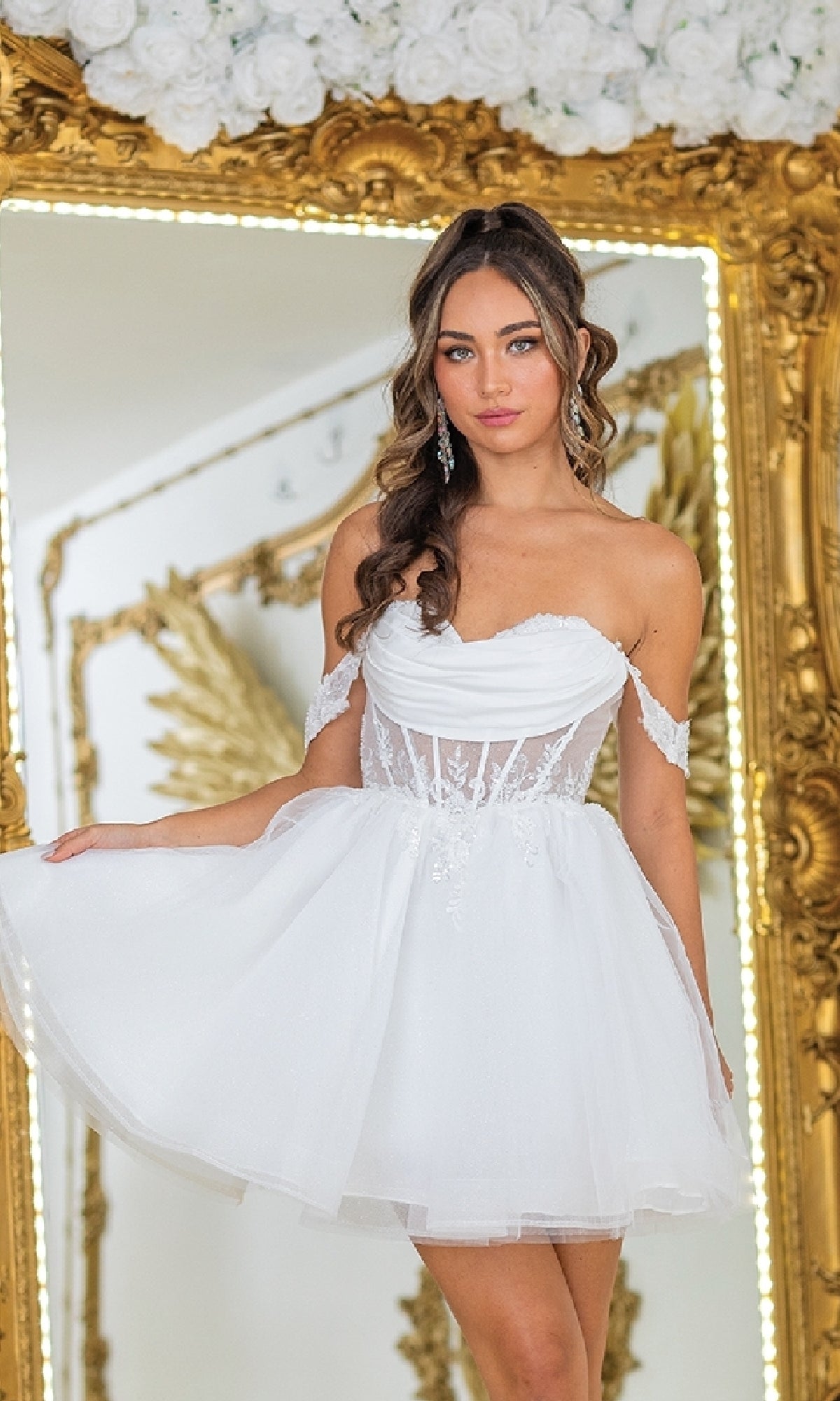 A woman in the Dancing Queen 3409 short babydoll prom dress stands before a large ornate gold mirror decorated with white flowers.