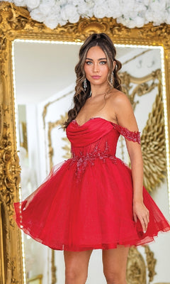 A woman in the Short Babydoll Prom Dress: Dancing Queen 3409, featuring off-the-shoulder floral details, stands before an ornate gold-framed mirror.