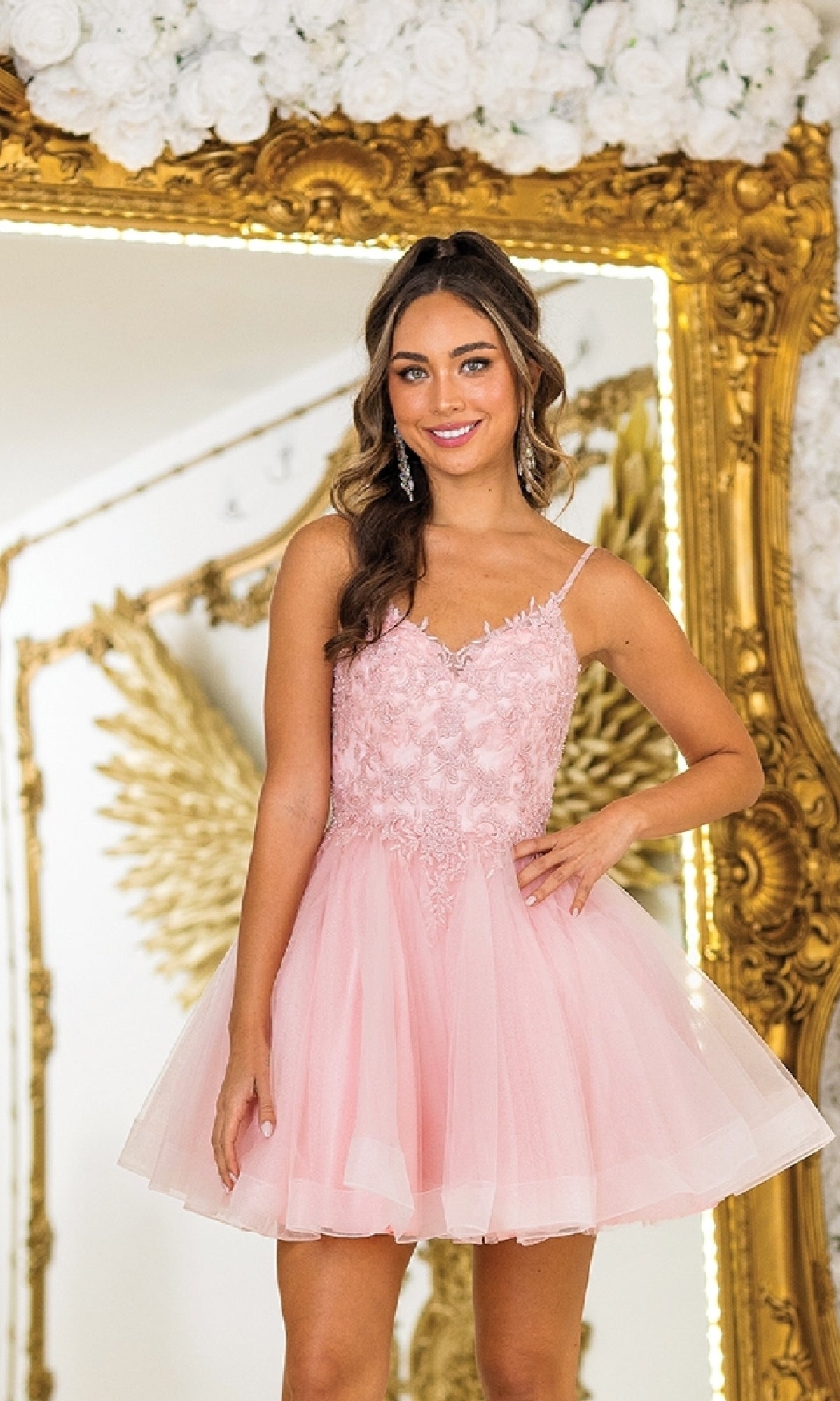 A woman in the Short Glitter-Tulle Prom Dress: Dancing Queen 3413 stands before a gold ornate mirror and white floral arrangement, smiling at the camera.