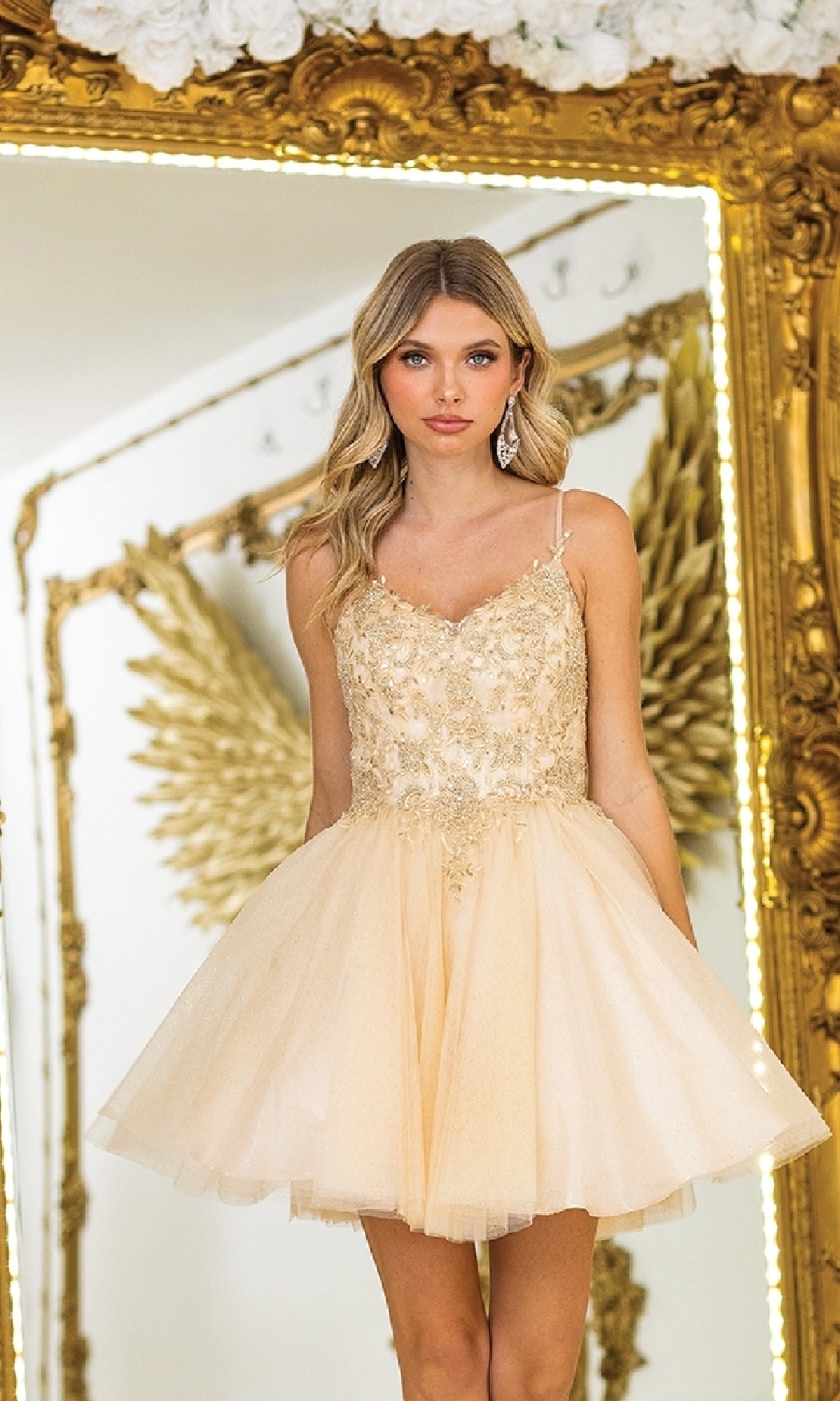 Wearing the Dancing Queen 3413 Short Glitter-Tulle Prom Dress, a woman stands before a decorative mirror framed with gold angel wings, her light gold embellished dress sparkling beautifully in the light.