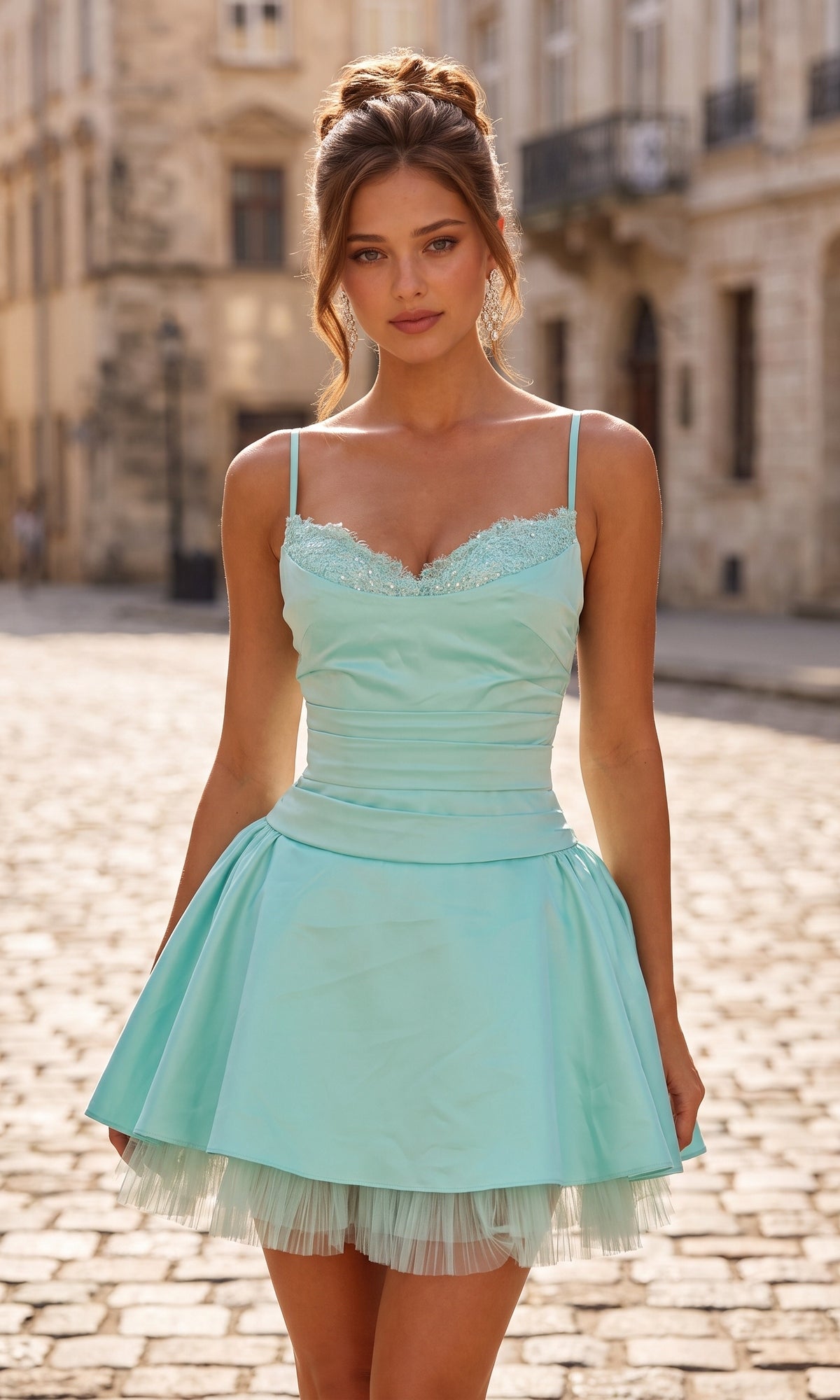 A woman with brown hair in an updo is wearing the La Femme 34144 short homecoming dress in aqua, featuring a fit-and-flare silhouette and satin bodice, as she stands on a cobblestone street lined with historic buildings in daylight.