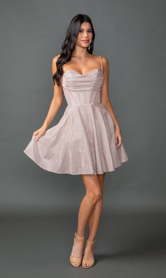 A woman poses against a gray background wearing the Short Prom Dress: Dancing Queen 3421—a light pink, sleeveless, knee-length dress with a corset bodice and spaghetti straps—paired with nude high-heeled sandals.