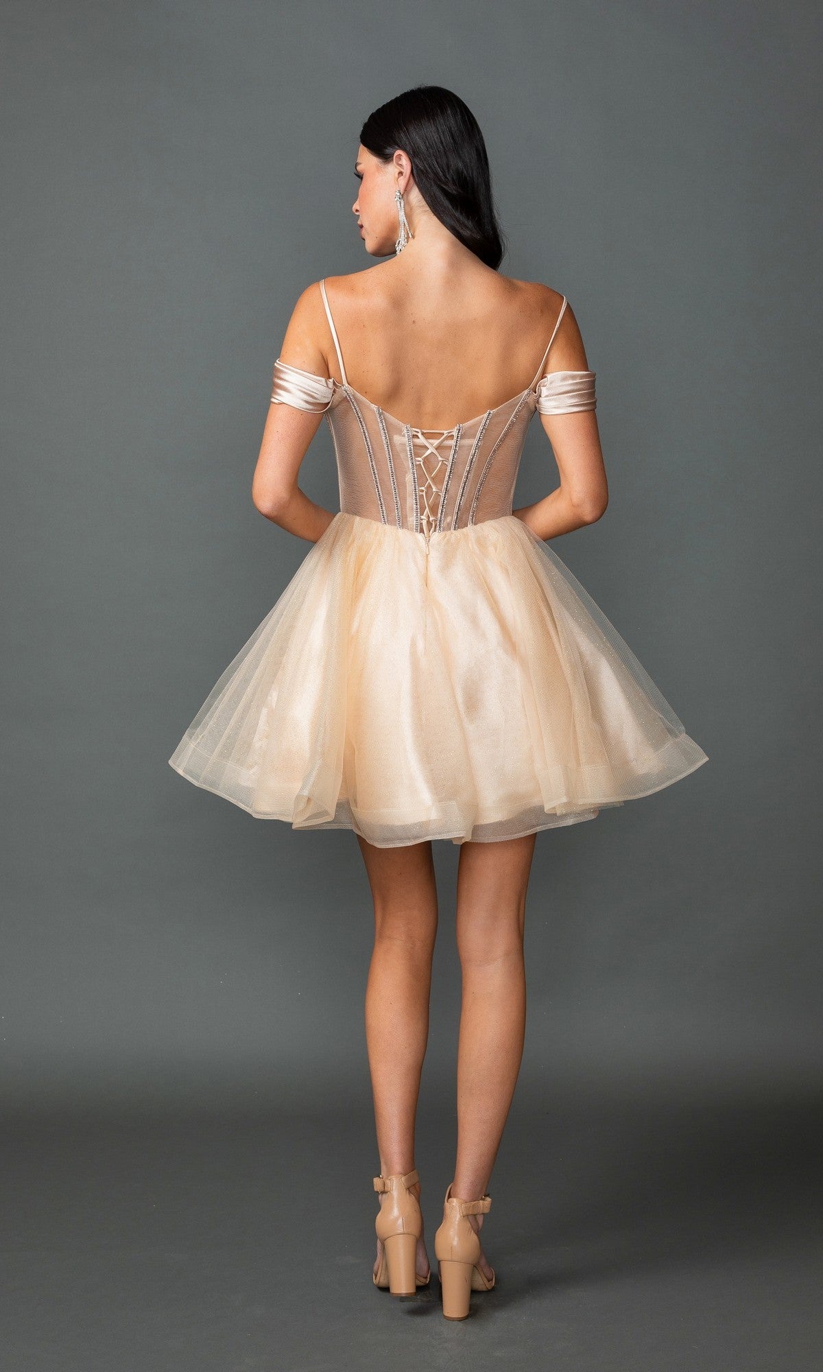 A woman poses against a gray background wearing the Dancing Queen 3428 short prom dress, featuring an off-the-shoulder design, tulle skirt, lace-up corset back, and high-heeled sandals.