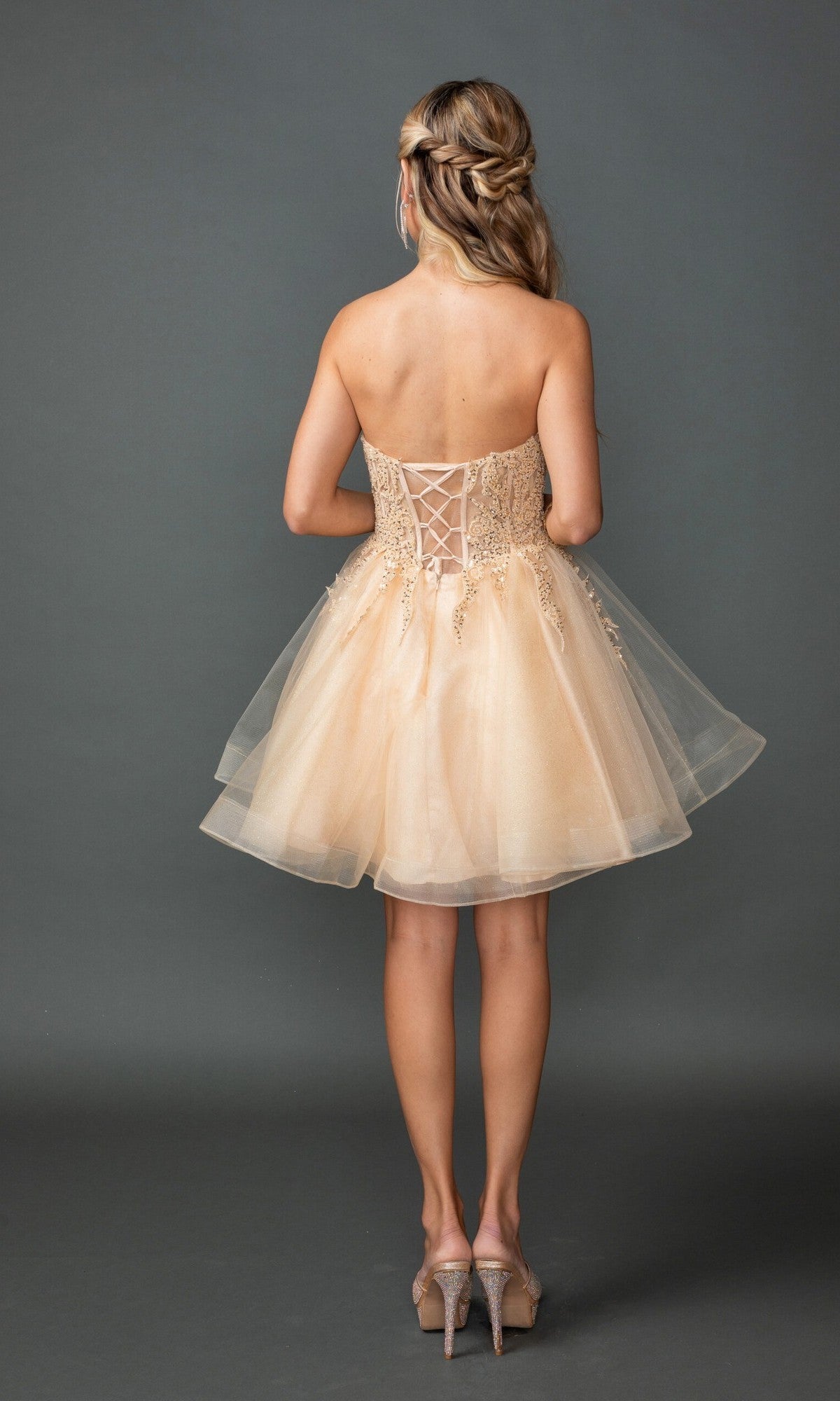 A woman stands facing away, wearing the Dancing Queen 3430 short homecoming dress featuring a strapless sweetheart neckline, beige tulle fabric, lace-up back, and paired with high heels against a plain gray background.