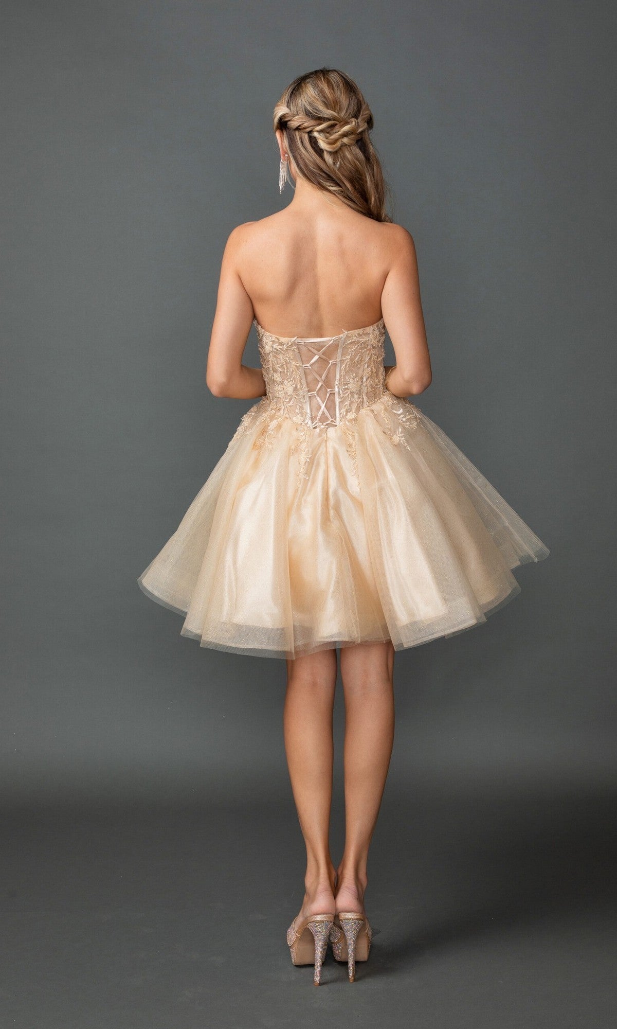 A woman faces away, showcasing the Dancing Queen 3431 short homecoming dress. This strapless design features an embroidered corset bodice and a lace-up back, complemented by a playful A-line skirt with tulle layers in shimmering gold. Her hair is styled in loose waves, and she completes the look with high-heeled shoes.