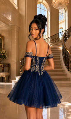 Wearing the Dancing Queen 3432 short homecoming dress—a navy blue, off-shoulder style with a boned corset, a-line skirt, and gold embroidery—a woman stands indoors near a grand staircase and large windows.