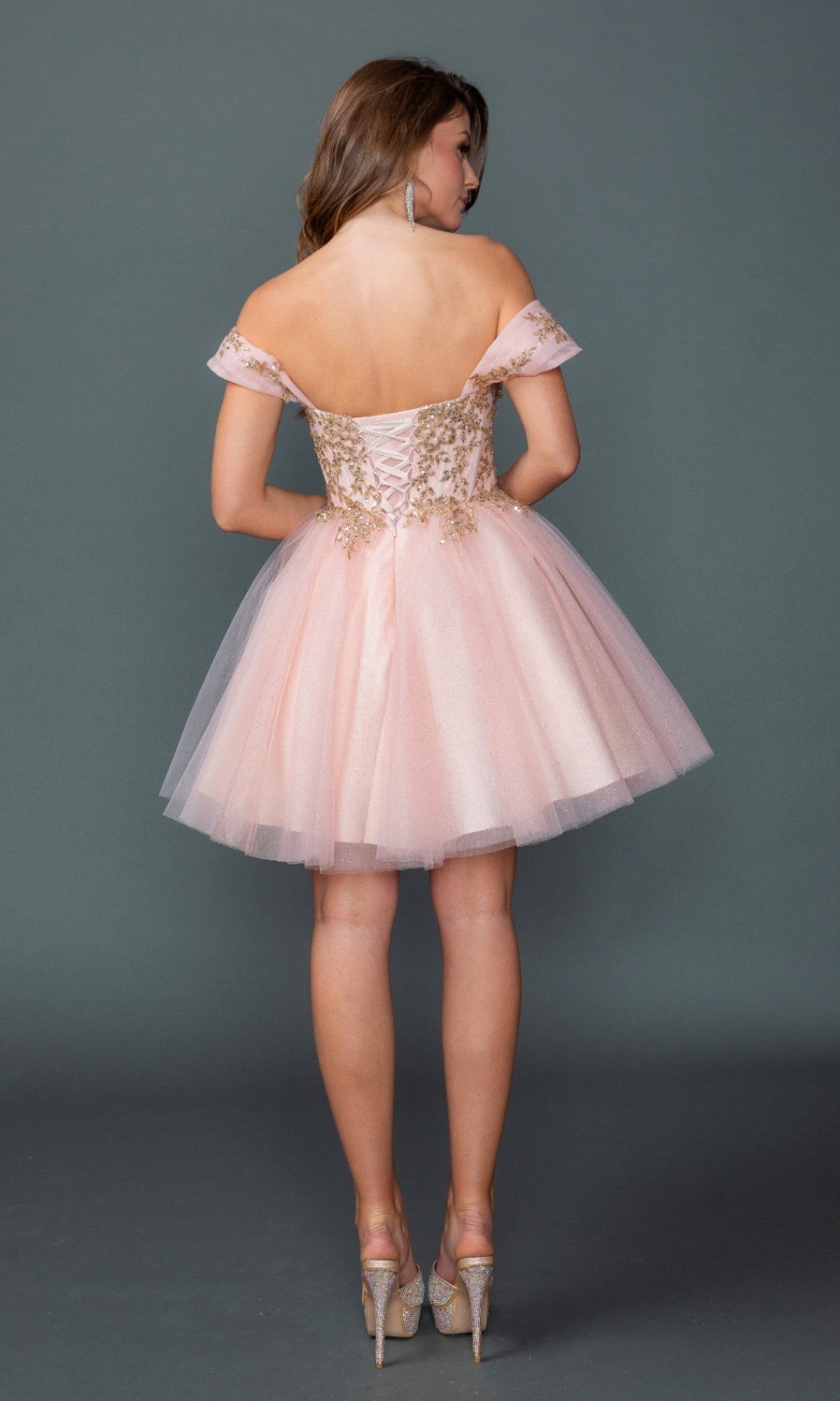 A woman stands with her back to the camera in the Dancing Queen 3433 short homecoming dress, showcasing its off-the-shoulder design, gold detailing, and embroidered corset bodice, paired with high heels against a plain gray background.