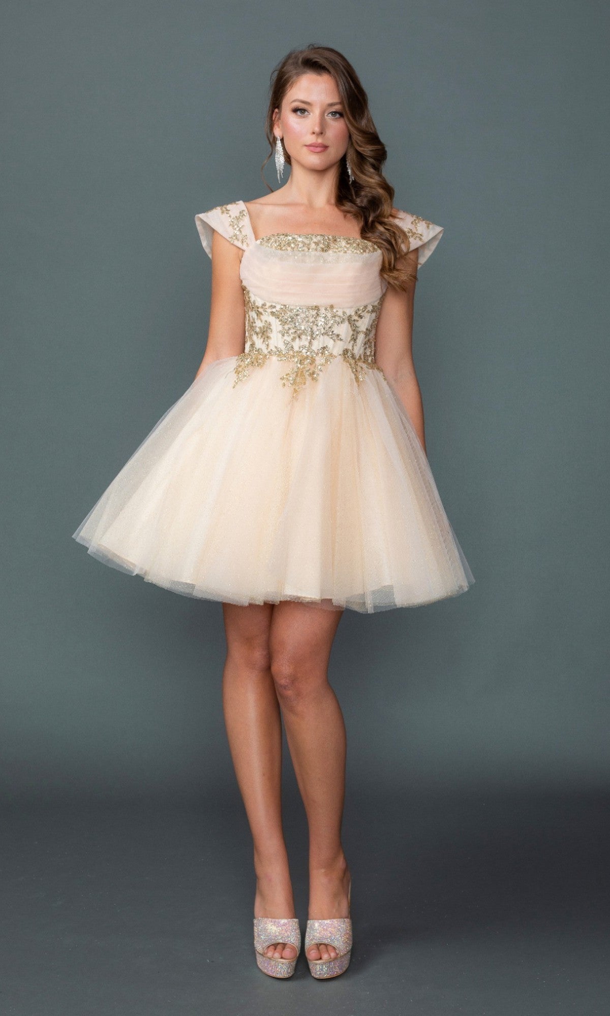 A woman stands against a plain gray background wearing the Dancing Queen 3433 short homecoming dress in champagne, featuring floral embroidery, an elegantly embroidered corset bodice with cap sleeves, and a delicate glitter tulle skirt. She completes the look with silver peep-toe heels.