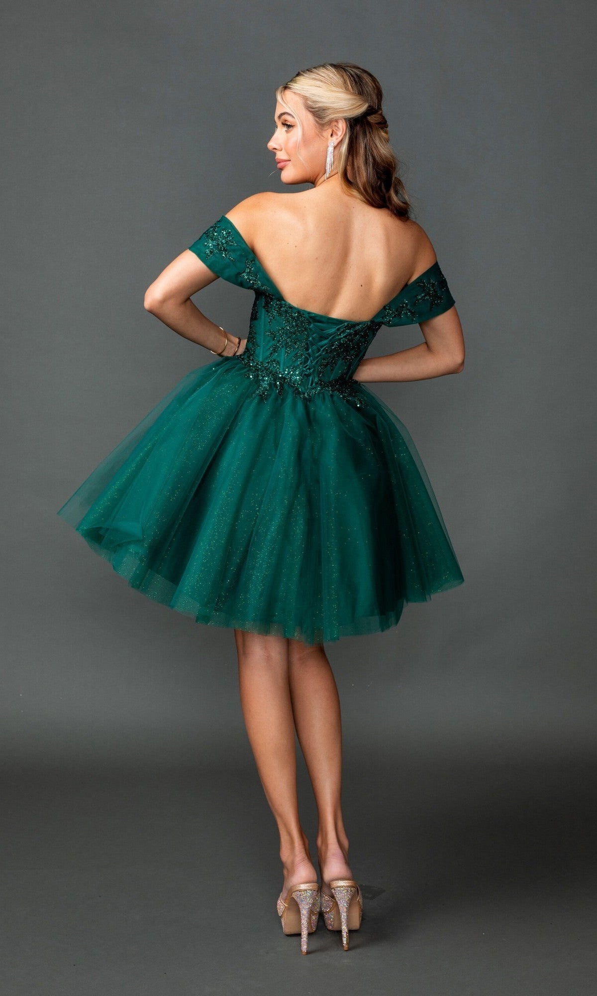 A woman wearing the Short Homecoming Dress: Dancing Queen 3433, which features an off-the-shoulder design, a short green glitter tulle skirt, and jeweled heels, stands with her back to the camera against a gray background.