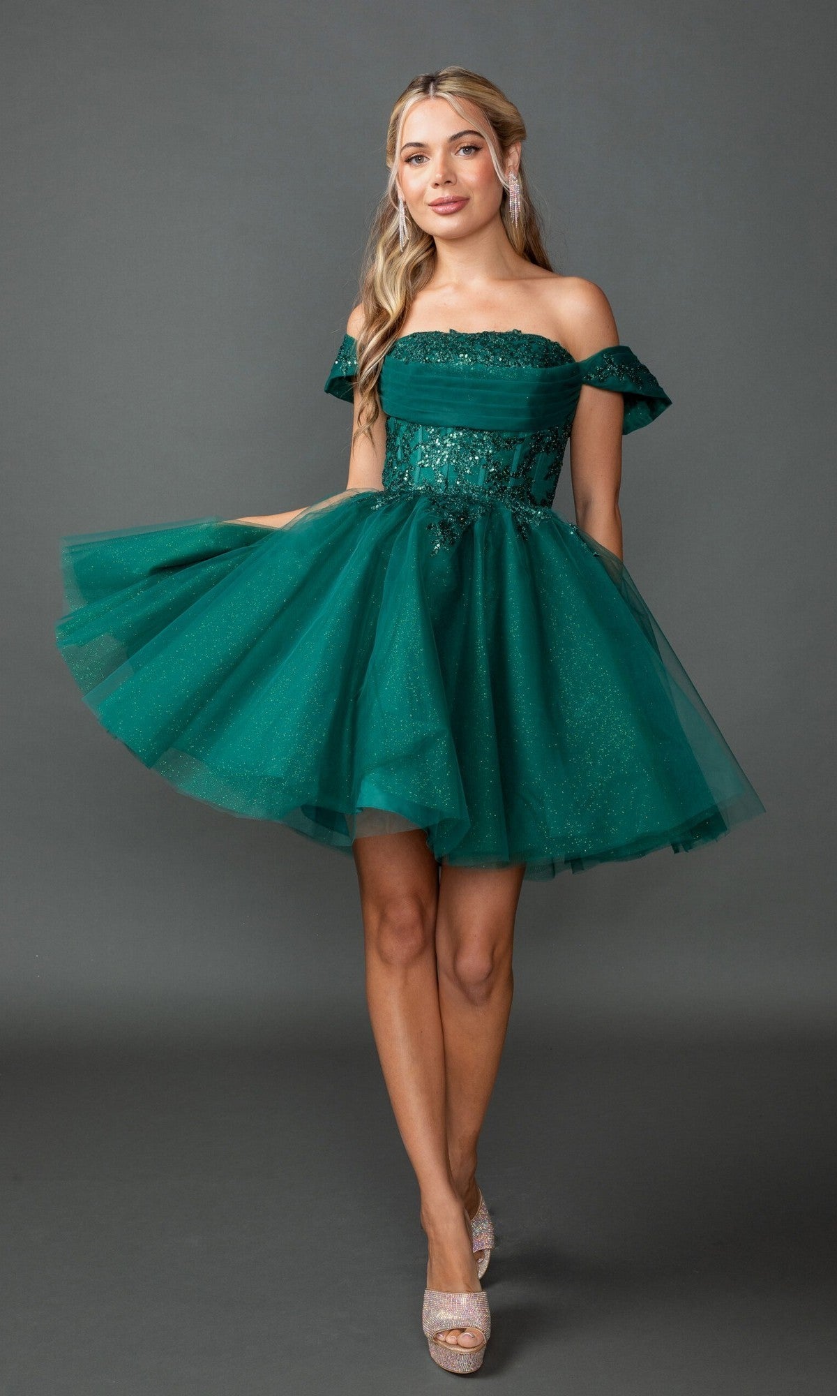 A woman showcases the Dancing Queen 3433 short homecoming dress in hunter green, featuring an off-the-shoulder design with a glitter tulle skirt and elegant sequin accents, complemented by an embroidered corset bodice, while standing against a gray background.