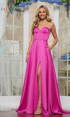 A woman stands indoors wearing the Strapless Corset Prom Dress: Colors Dress 3434 in bright pink, featuring a front slit and side pockets, posing before glass doors decorated with blue flowers.
