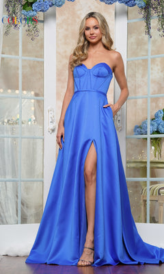 A woman with long, wavy blonde hair poses indoors in front of flower-decorated glass doors, wearing the Strapless Corset Prom Dress: Colors Dress 3434 in bright blue with a thigh-high slit.