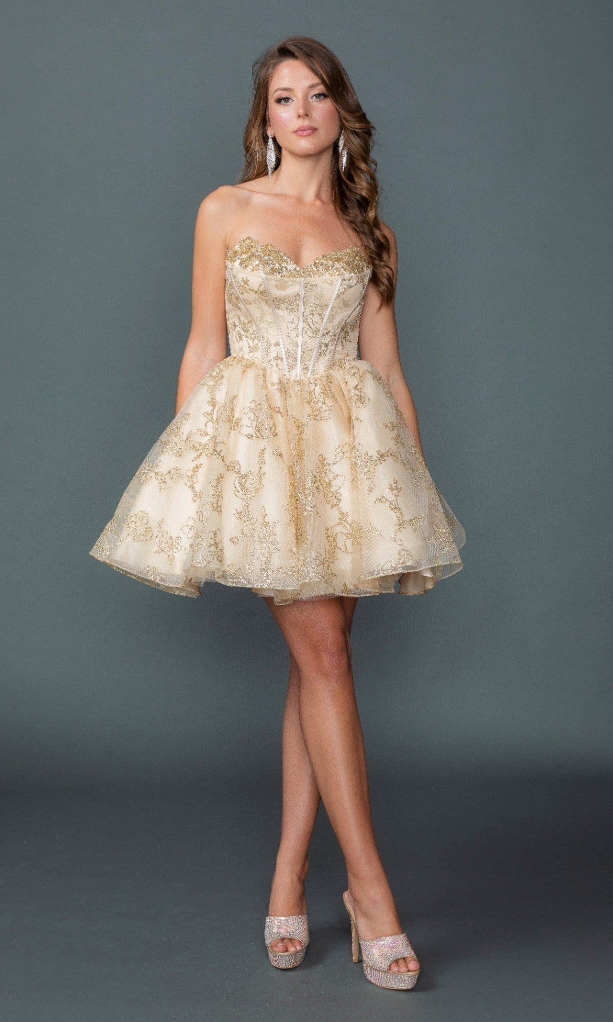 A woman stands against a gray backdrop wearing the Short Homecoming Dress: Dancing Queen 3436 in champagne, featuring a strapless sweetheart neckline, lace details, and a short babydoll silhouette, styled with sparkling high heels and drop earrings.