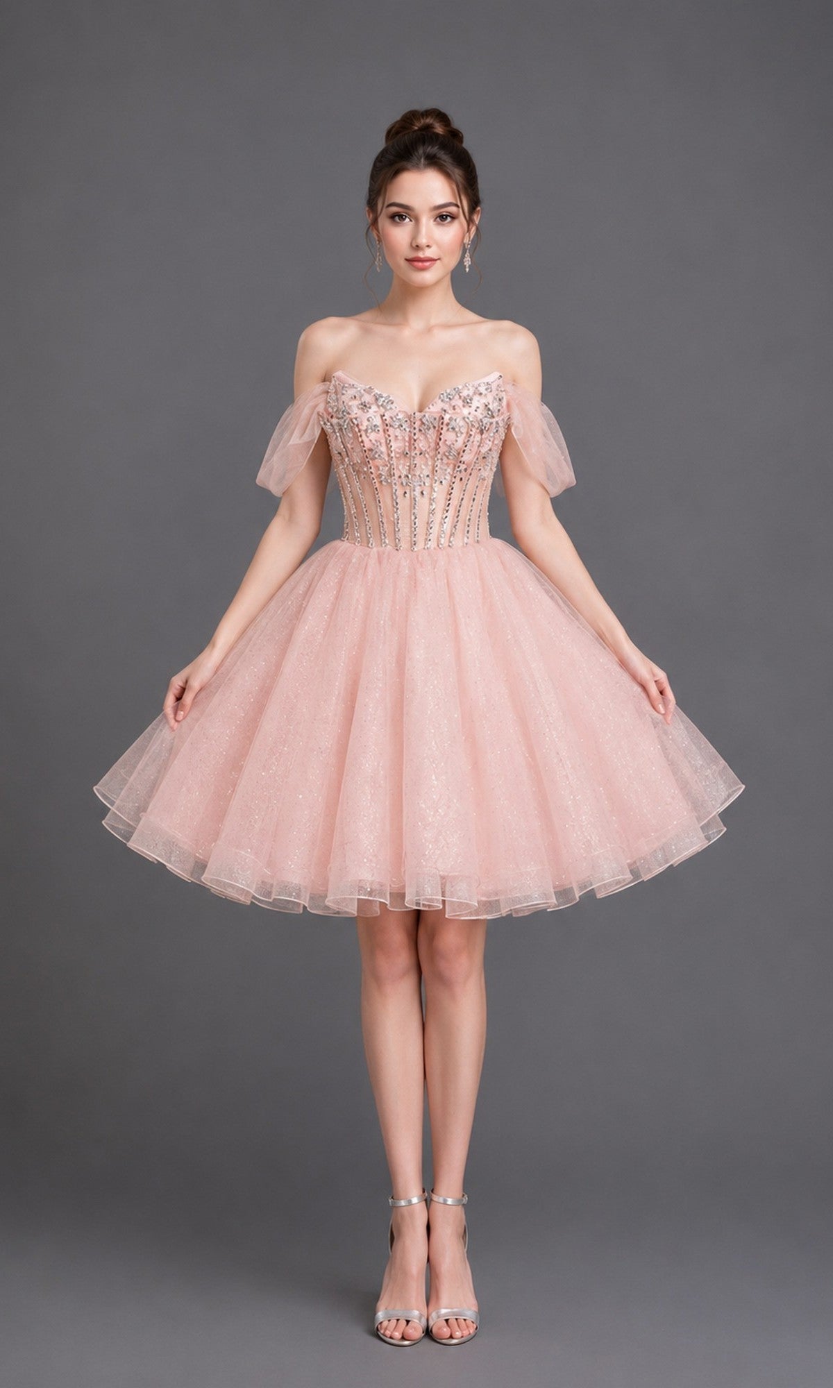A woman stands against a gray background wearing the Dancing Queen 3438 short homecoming dress in pink, featuring off-the-shoulder straps, a fitted embellished bodice, and a sparkly tulle skirt, paired with strappy silver heels.