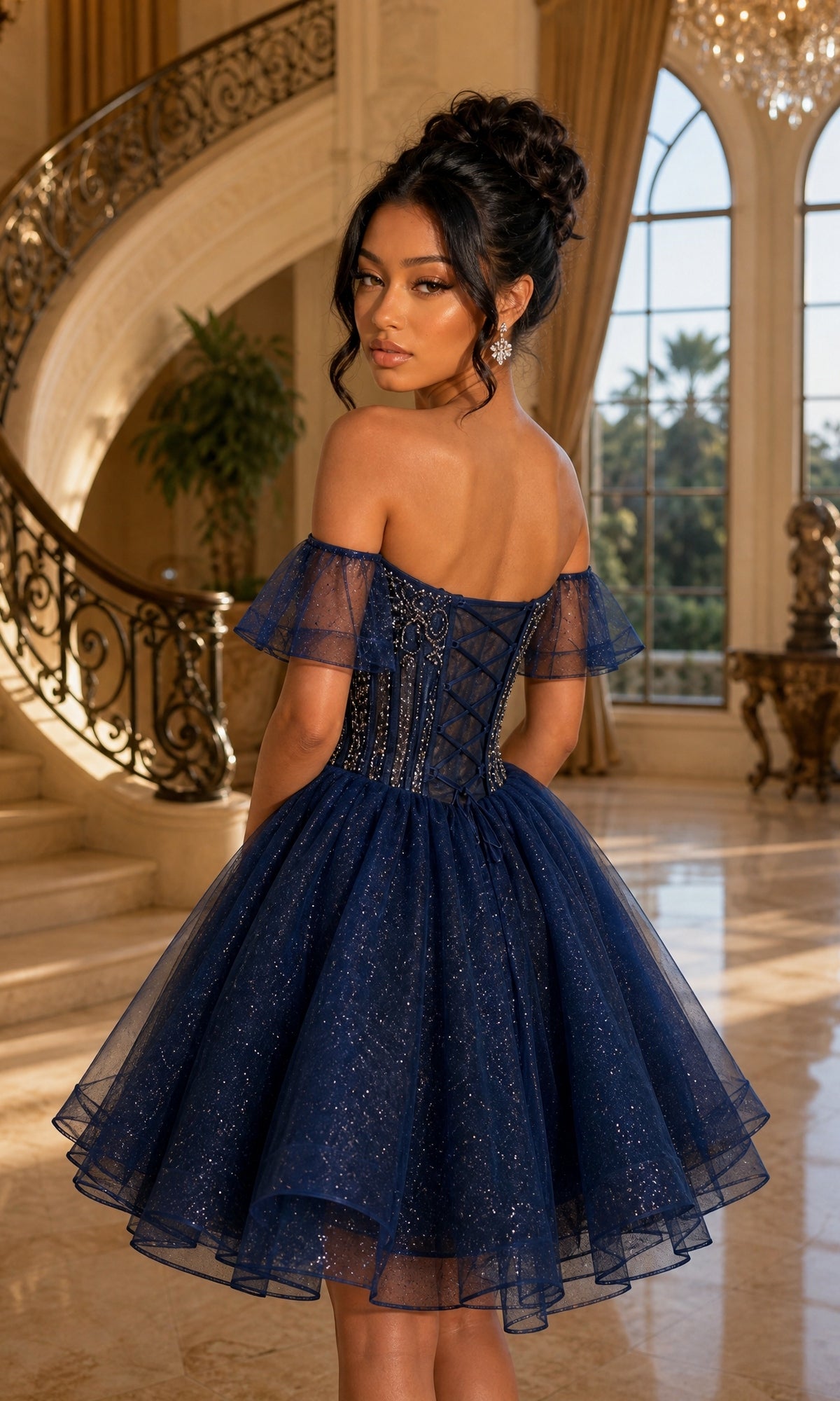 A woman in the elegant “Short Homecoming Dress: Dancing Queen 3439,” featuring an off-the-shoulder navy blue design with a sheer glitter tulle overlay, stands indoors near a curved staircase and large windows.