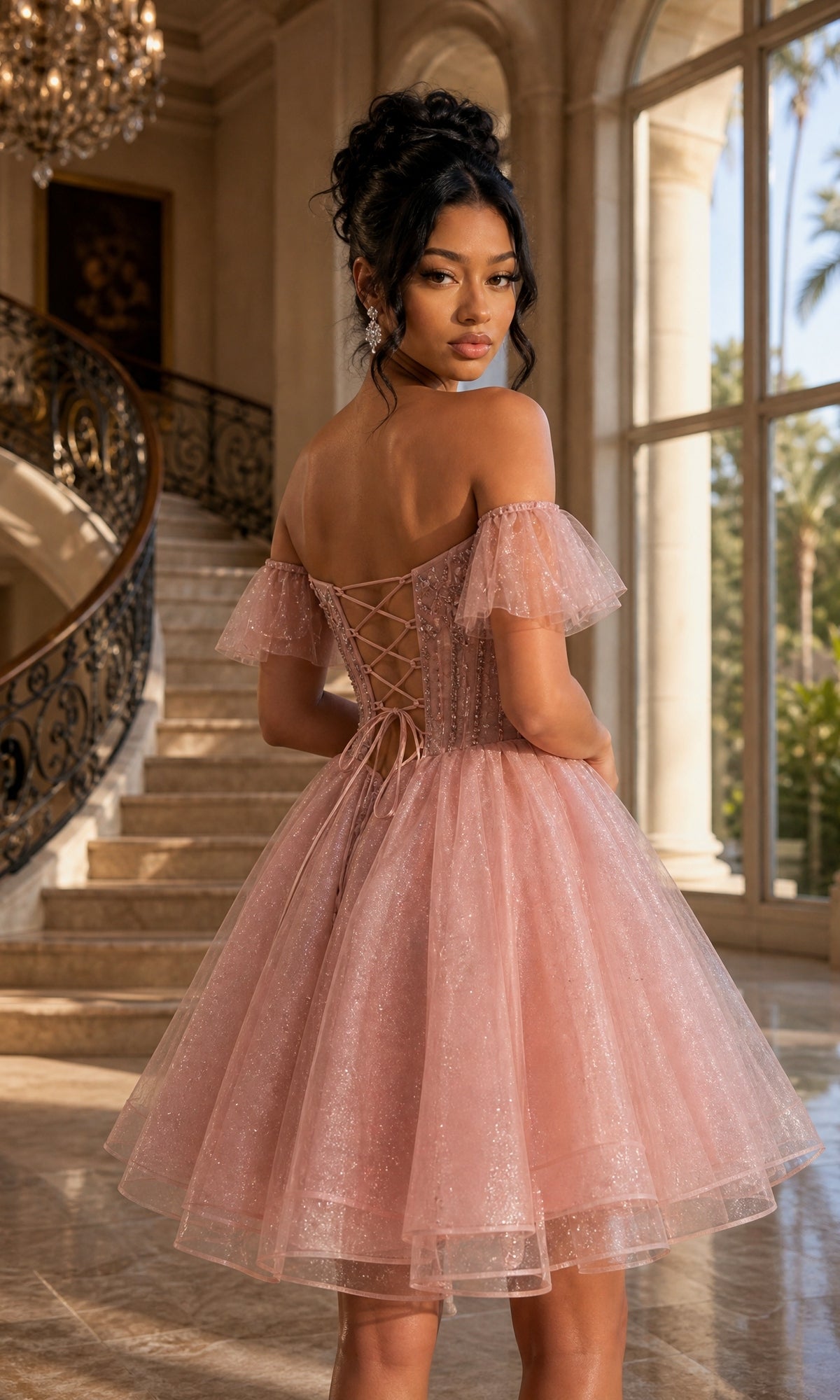A woman stands indoors near a staircase and large windows, wearing the Dancing Queen 3439—a short, off-the-shoulder pink homecoming dress featuring a lace-up back.