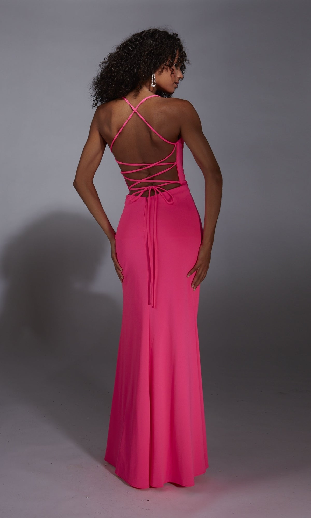A woman wearing the Aerie Couture 343 long prom dress in bright pink, featuring an open back and lace-up corset bodice, stands facing away from the camera.