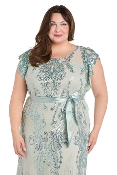 A woman stands smiling against a white background, wearing the Plus-Size Sage Green Formal Dress: R&M Richards 3465W, featuring sequin print, short sleeves, and a satin waist tie.