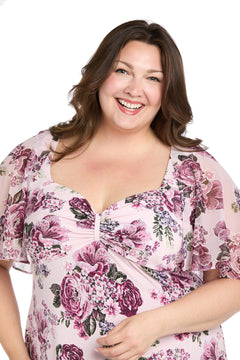 A woman with long brown hair smiles, wearing the R&M Richards 3467W plus-size pink party dress with floral pattern and sheer short sleeves, standing against a white background.