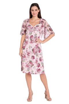 A woman smiles while wearing the Modest Pink Print Party Dress (R&M Richards 3467), featuring a floral print and matching sheer short-sleeved overlay, paired with neutral heels.