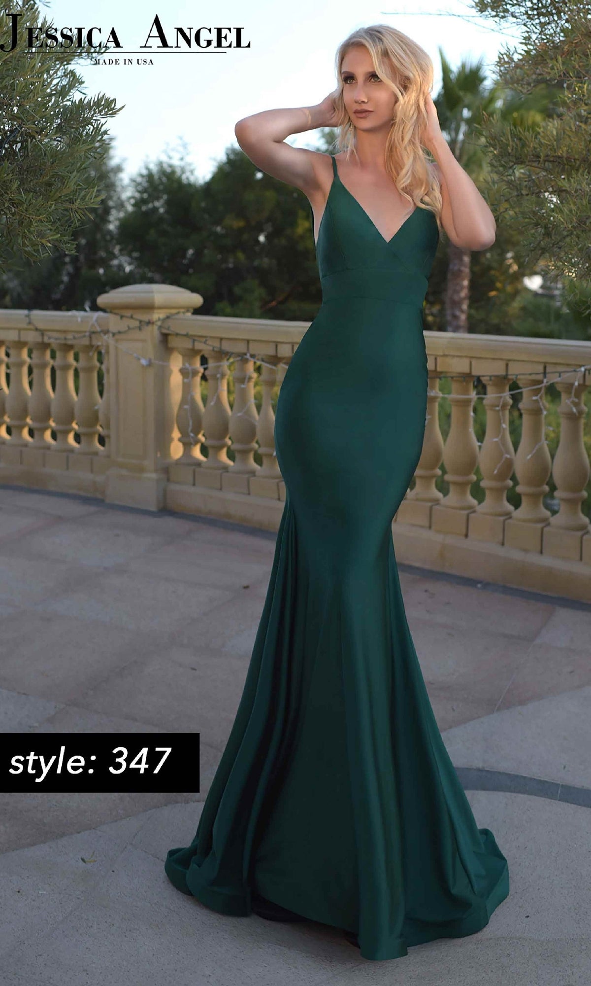 A woman stands outdoors in a sleeveless, floor-length emerald green Open-Back Long Prom Dress: Jessica Angel 347 with a fitted bodice and flared skirt. The text 