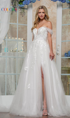 A woman stands before glass doors, exuding elegance in the Long Glitter Prom Ball Gown: Colors Dress 3474—an off-the-shoulder white gown with floral embroidery and a thigh-high slit.