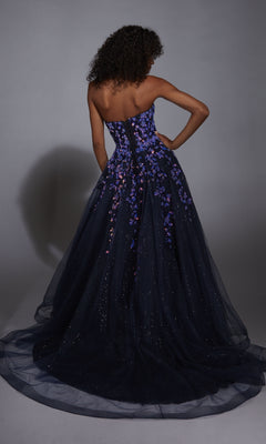 A woman faces away from the camera in the Aerie Couture 350 navy blue prom ball gown, featuring a strapless fitted bodice with purple floral appliques and a voluminous, glittery A-line tulle skirt.