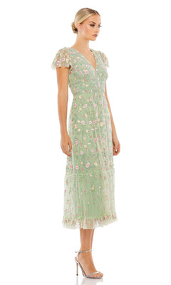 Woman in the Mac Duggal 35104 embroidered wedding-guest dress with floral details, light green hue, ruffled hem, and short sleeves, stands in profile wearing silver high-heeled sandals against a white background.