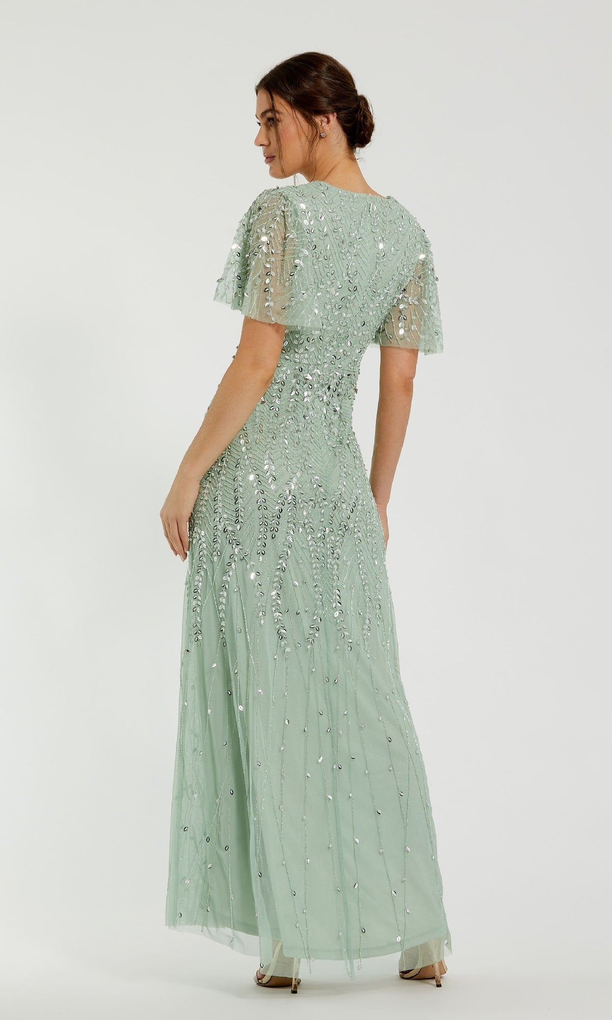 A woman faces away in the Mac Duggal 35109 Flutter-Sleeve Long Formal Dress—a floor-length, light green gown with short flutter sleeves and sequin embellishments—set against a plain white background.
