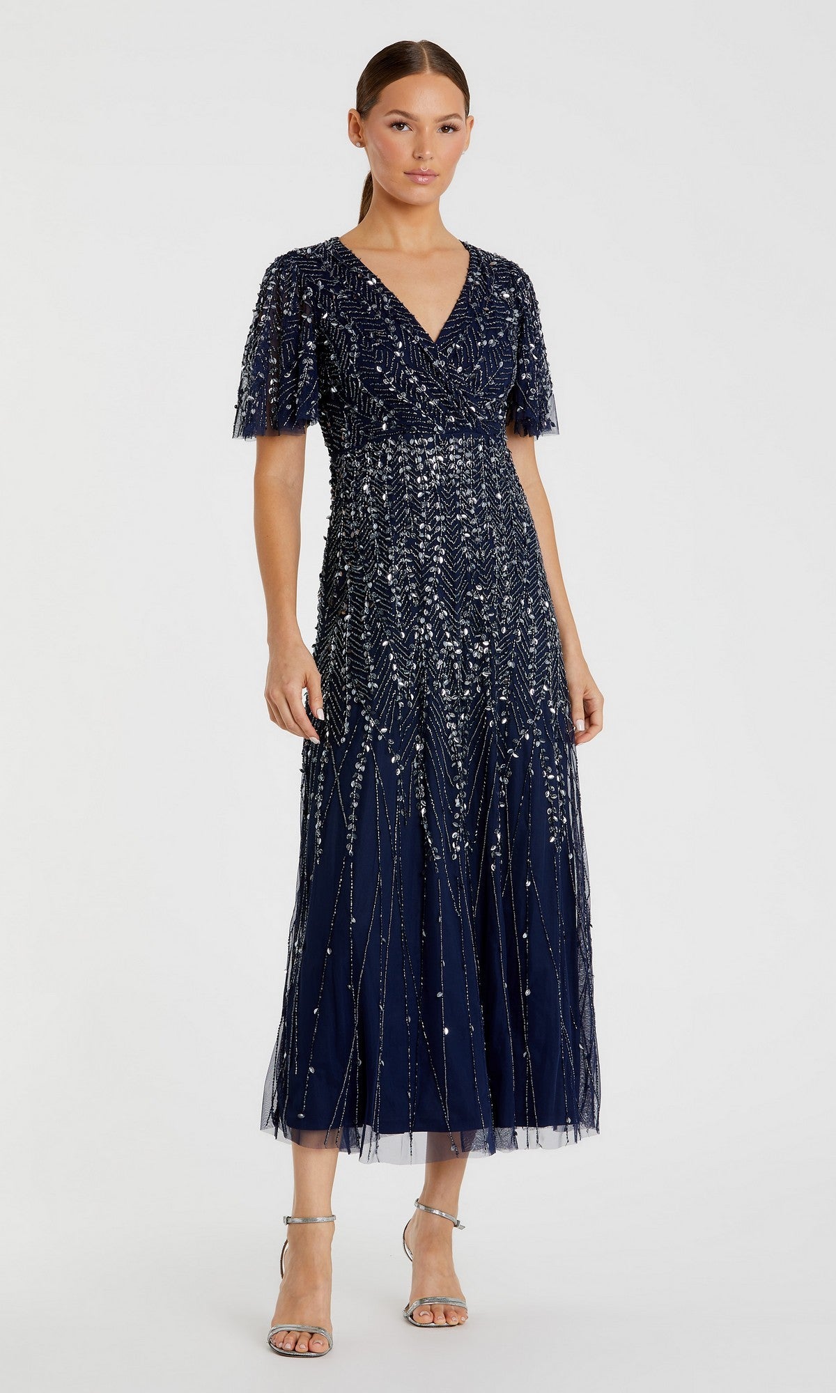 A woman wears the Mac Duggal 35116 beaded navy blue midi semi-formal dress with short sleeves and silver strappy heels, standing against a plain light background.