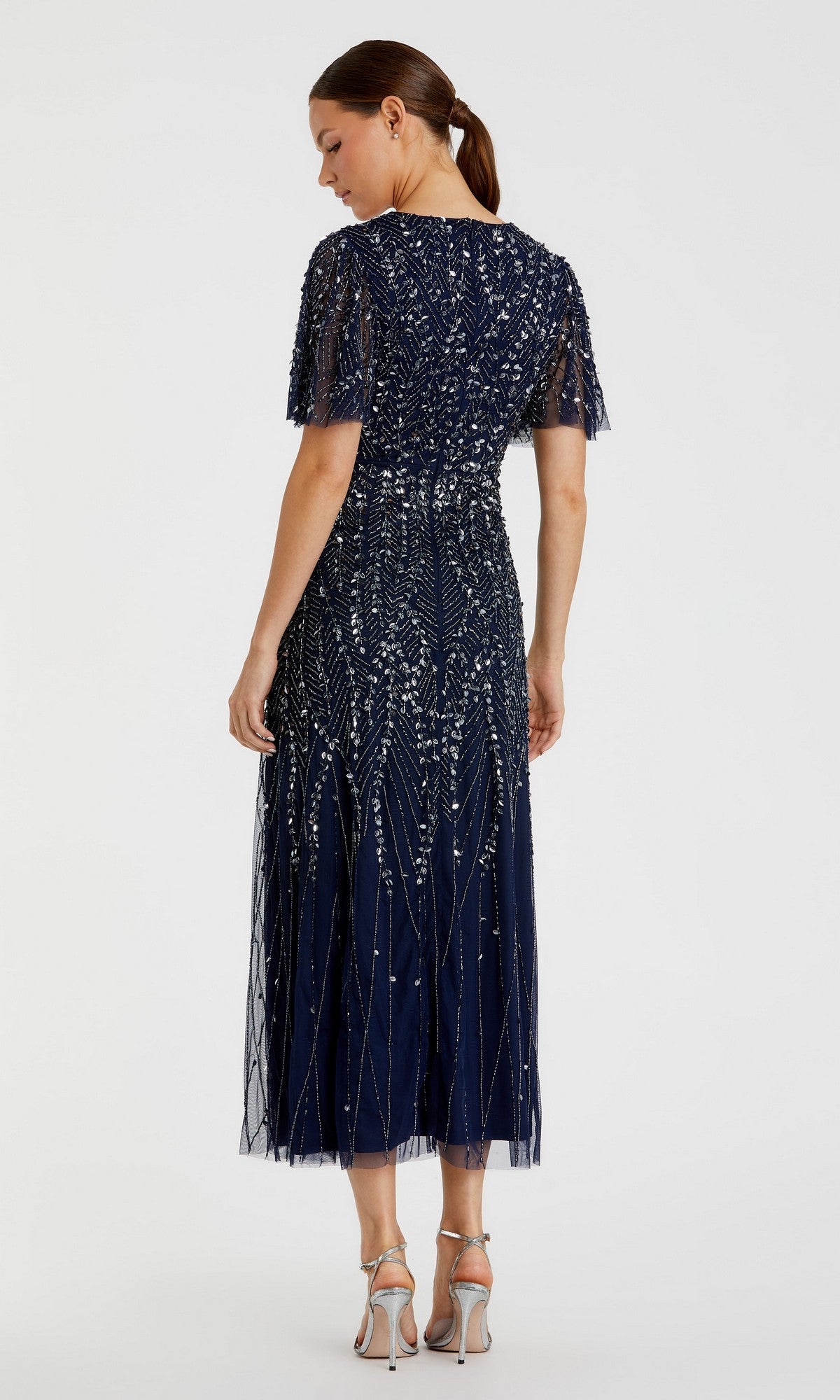 A woman stands facing away in the Beaded Midi Semi-Formal Dress: Mac Duggal 35116, featuring navy blue beads, silver embellishments, and short flutter sleeves. Her hair is tied back and she wears silver high-heeled sandals.