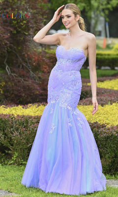 A woman smiles and touches her hair while standing on grass, wearing the Colors Dress 3529—an embroidered, strapless mermaid prom dress.