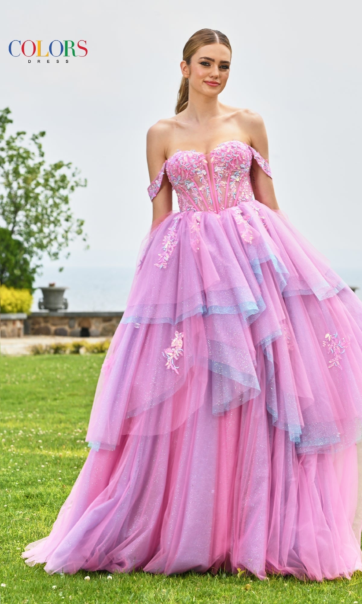 A woman stands outdoors on grass in the Off-Shoulder Long Prom Ball Gown: Colors Dress 3534, featuring floral embroidery and tulle. Cloudy skies, trees, and a stone wall create an enchanting backdrop.
